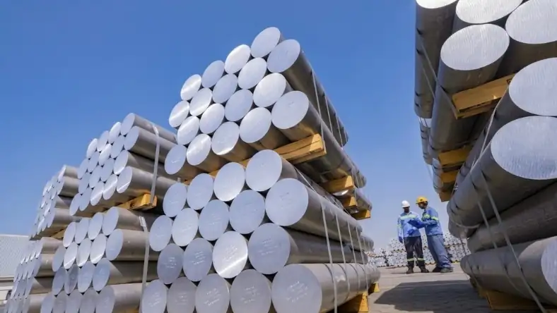 Stacked aluminum billets on pallets at an outdoor yard with two workers in safety gear inspecting the load