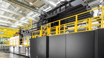 Heavy black industrial machine with yellow safety barriers and an overhead crane in a manufacturing facility